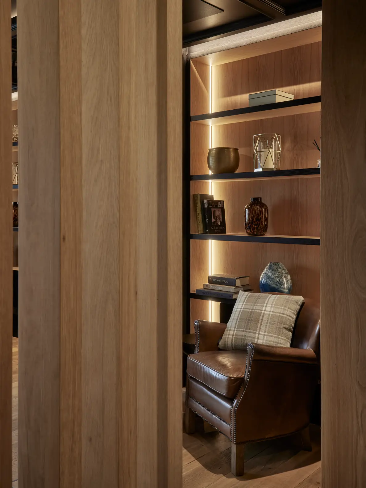 Brown leather lounge chair with plaid cushion set beside softly lit wooden shelving in Alderley Edge office.